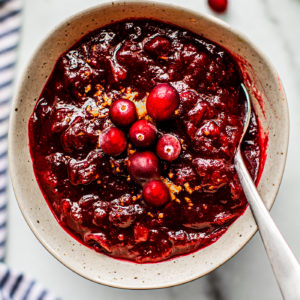 Speckled serving bowl full of cranberry sauce, topped with orange zest and fresh cranberries.