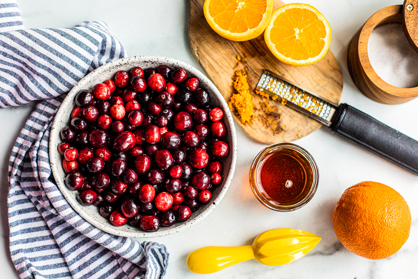 Bowl of fresh cranberries, a sliced orange with orange zest on a cutting board, a small jar or honey, and a salt crock.
