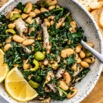 Serving bowl full of sauteed kale, white beans, sardines, and lemon wedges.
