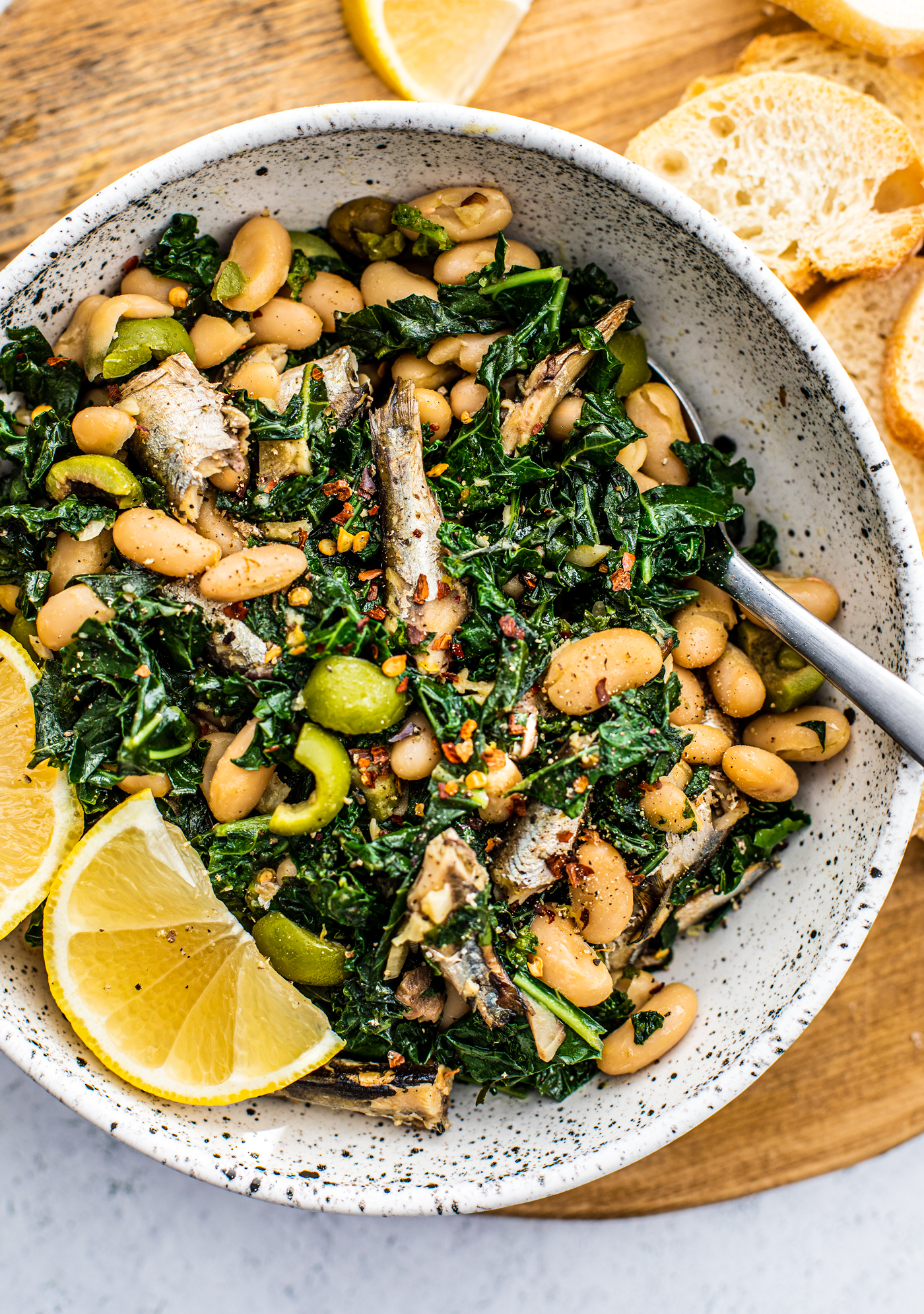 Serving bowl full of sauteed kale, white beans, sardines, and lemon wedges.