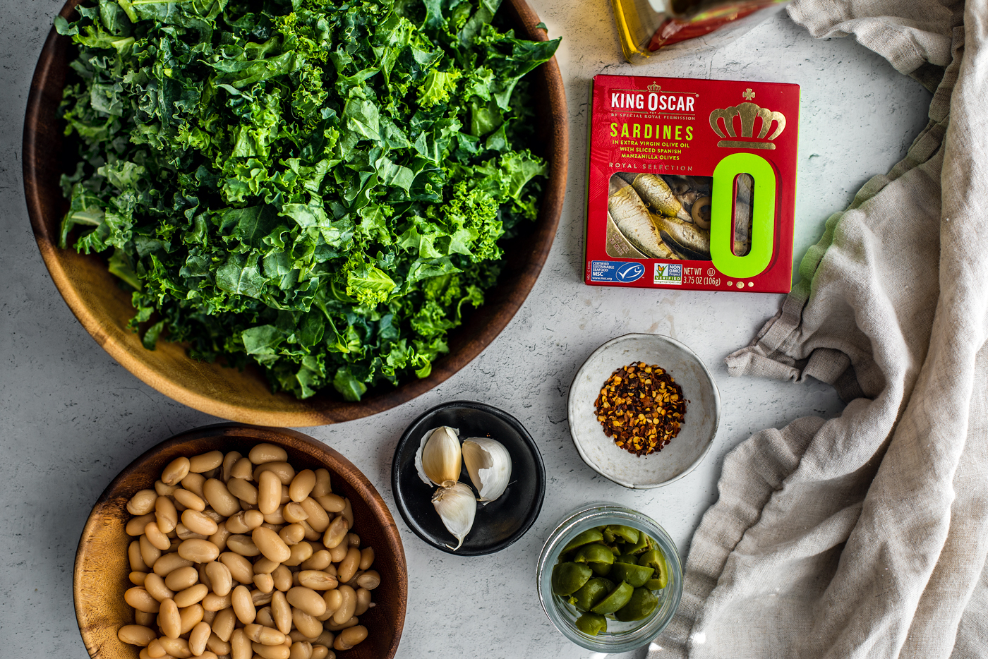 Big bowl of chopped kale, bowl of cannellini beans, garlic cloves, chili flakes, and olives next to a container of King Oscar sardines.