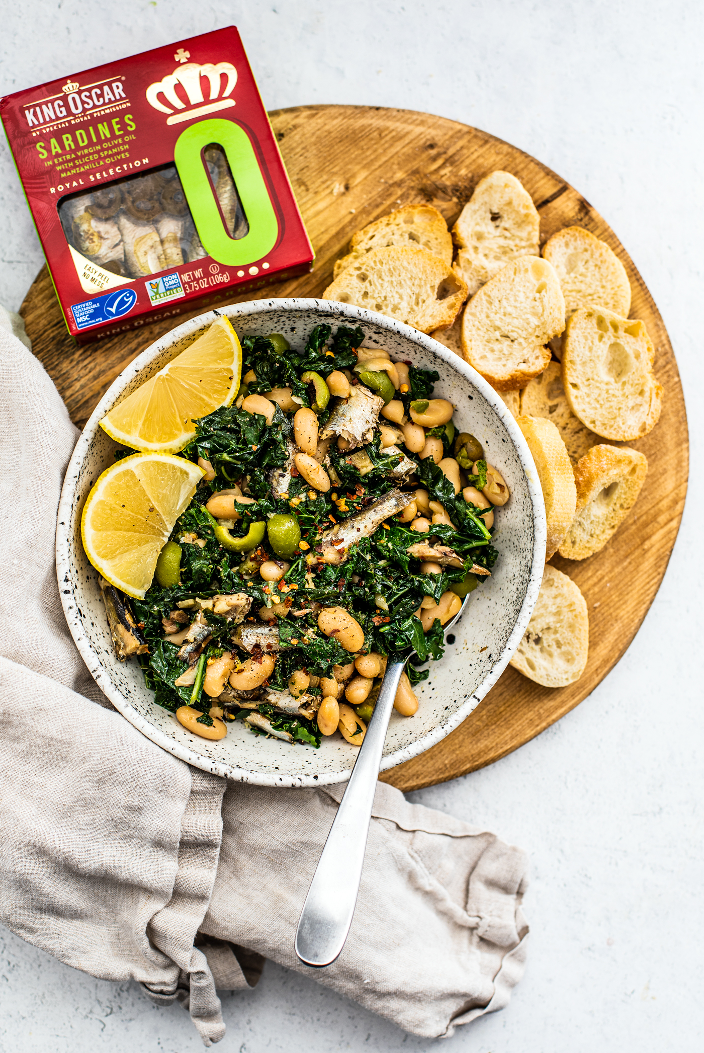 Serving board with bowl of greens and beans, toasted baguette slices, and a package of King Oscar sardines.