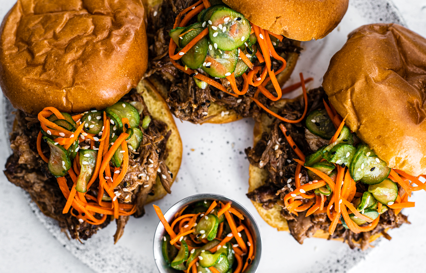 Serving plate with pulled pork sandwiches topped with pickled cucumbers and carrots.