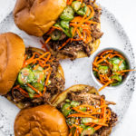 Serving plate with pulled pork sandwiches topped with pickled cucumbers and carrots.