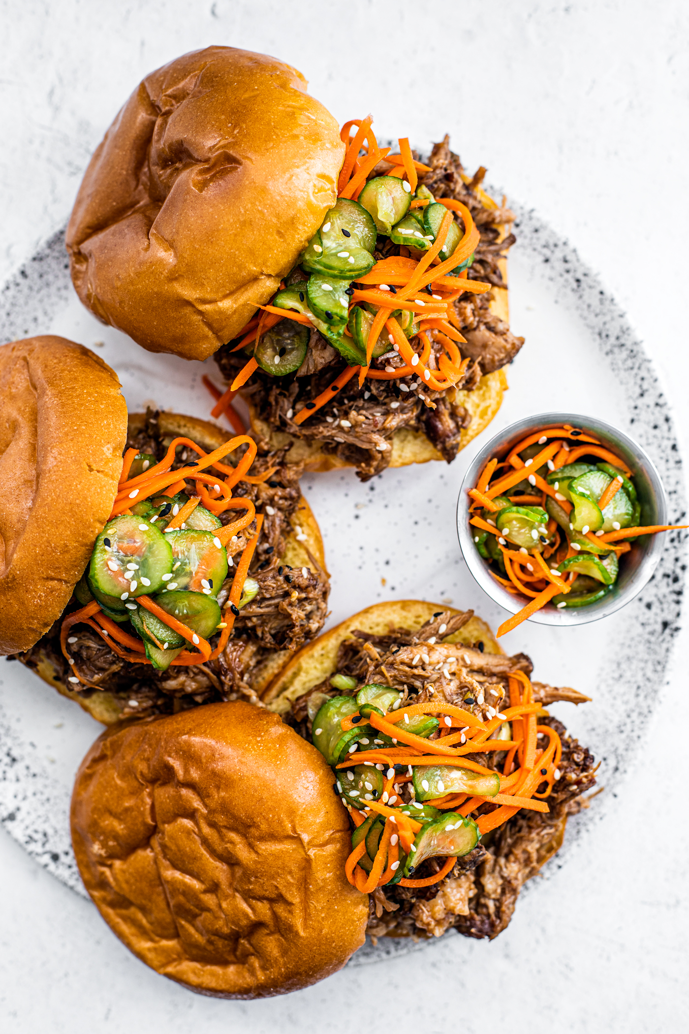Serving plate with pulled pork sandwiches topped with pickled cucumbers and carrots.