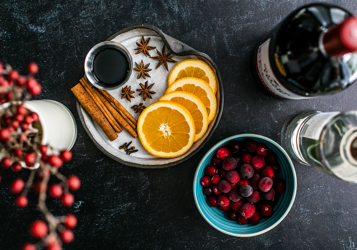 Plate with orange slices, cinnamon sticks, star anise, and dip bowl of maple syrup next to bowl of cranberries, bottle of red wine, and bottle of vodka.