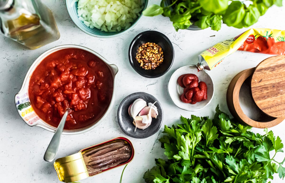 Flatlay of ingredients including bowl of crushed tomatoes, tin of anchovies, fresh herbs, and pinch bowls of garlic, chilis, and tomato paste.
