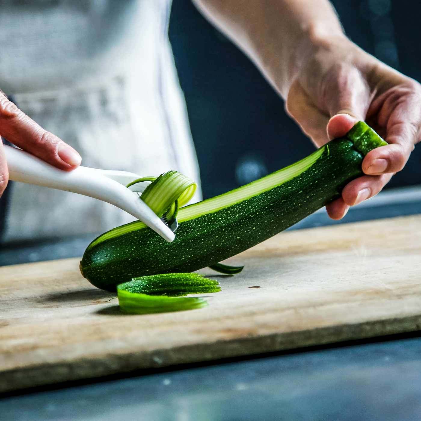 Hands peeling zucchini with a Y Peeler on a cutting board.