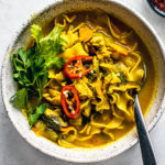 Bowl of curry chicken noodle soup garnished with fresh parsley and sliced chiles, and a small bowl of hot sauce next to it.