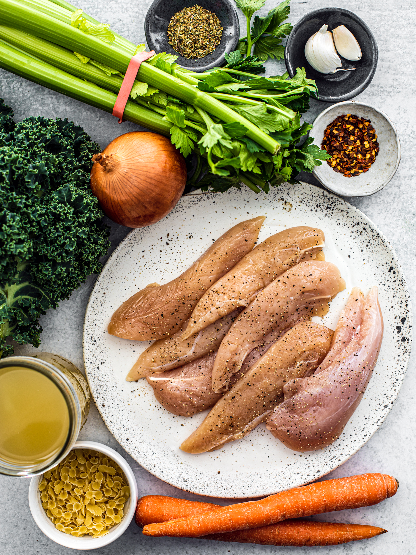 Uncooked chicken tenderloins on a plate surrounded by kale, celery, carrots, spices, broth, and dry pasta.