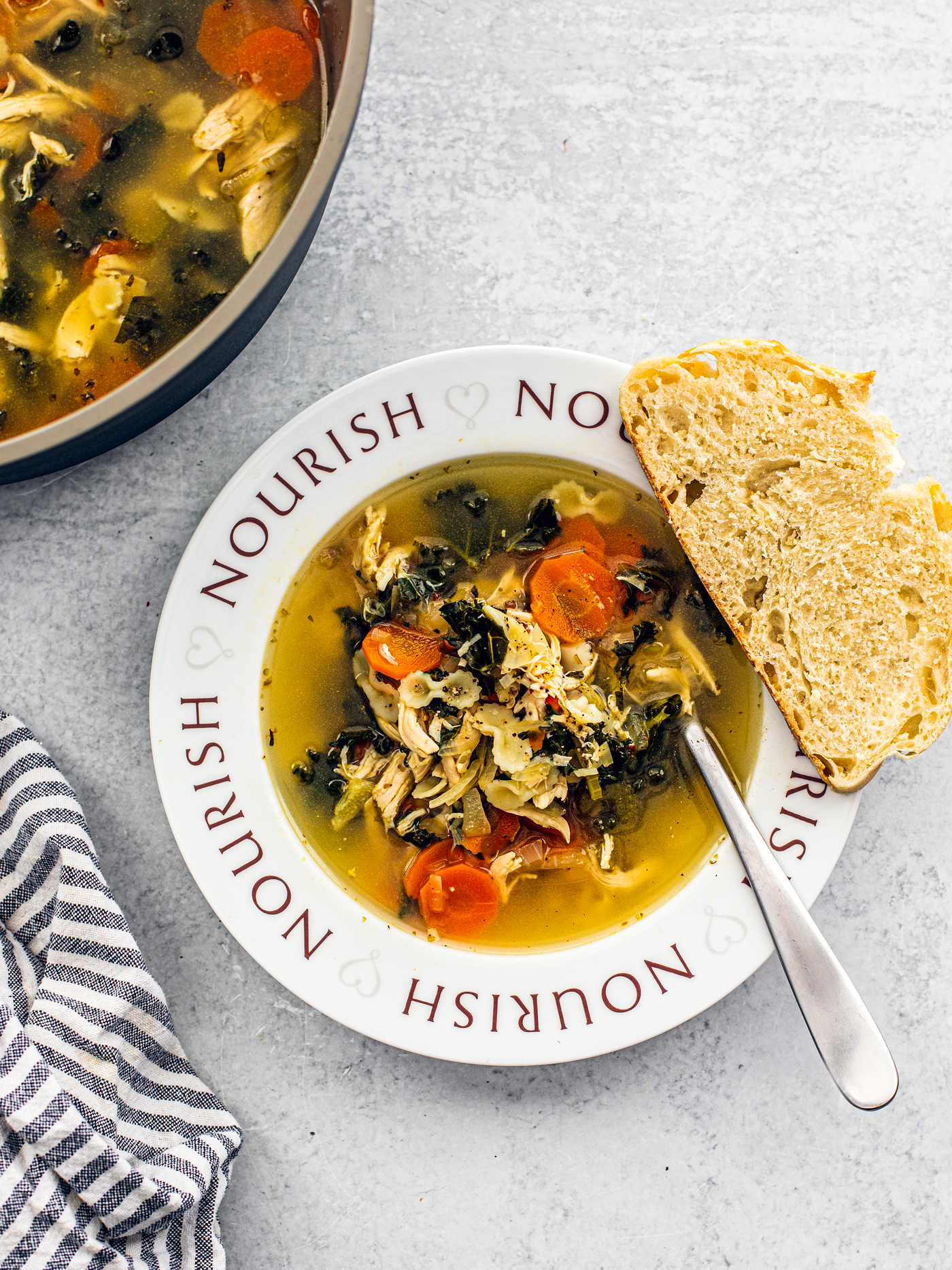 Soup bowl with the word "Nourish" written around it, filled with chicken soup next to a dish towel and pot of soup.