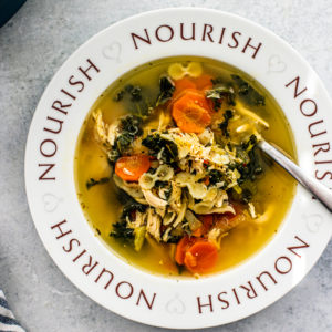 Soup bowl with the word "Nourish" written around it, filled with chicken soup next to a dish towel and pot of soup.