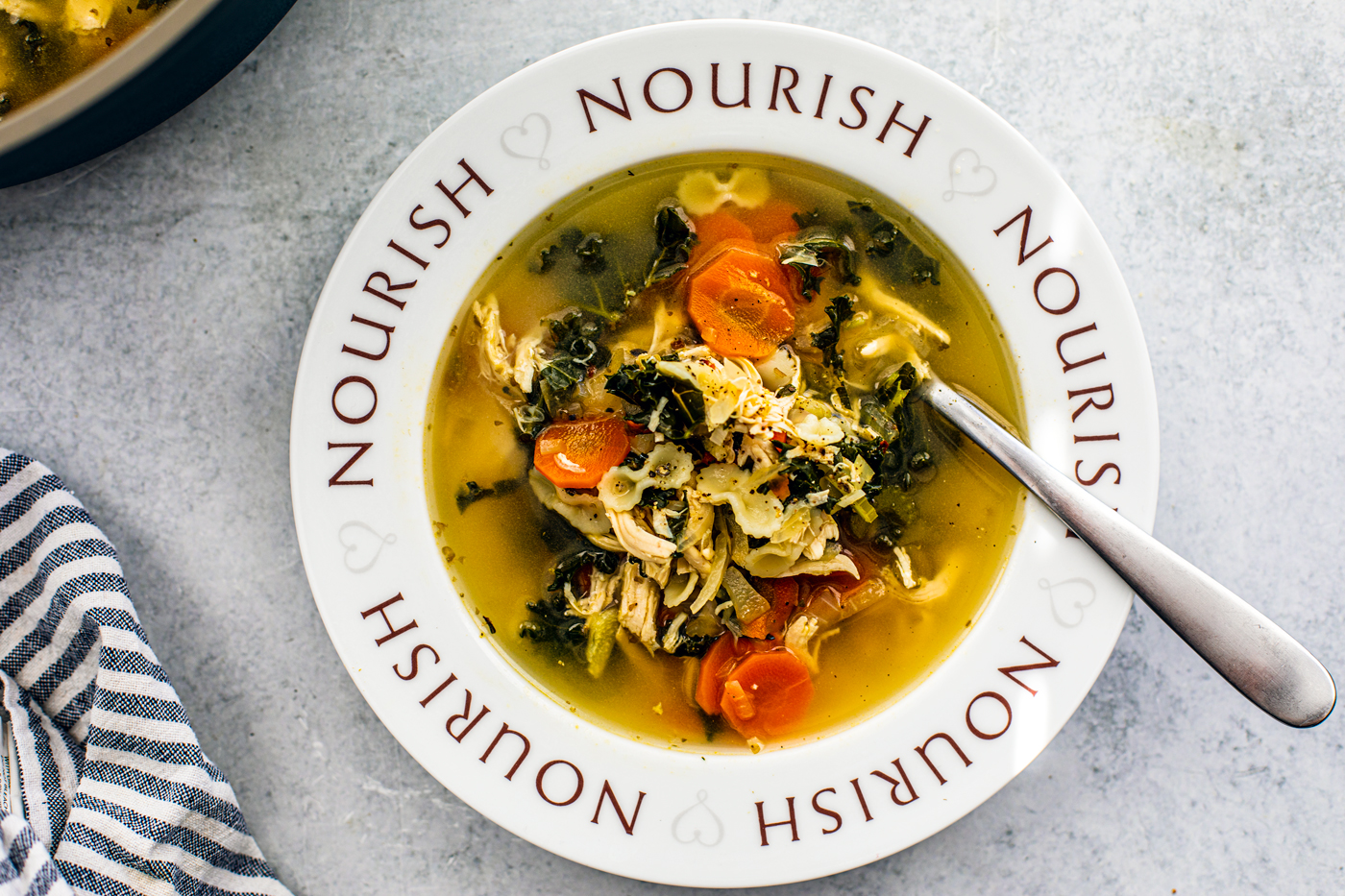 Soup bowl with the word "Nourish" written around it, filled with chicken soup next to a dish towel and pot of soup.