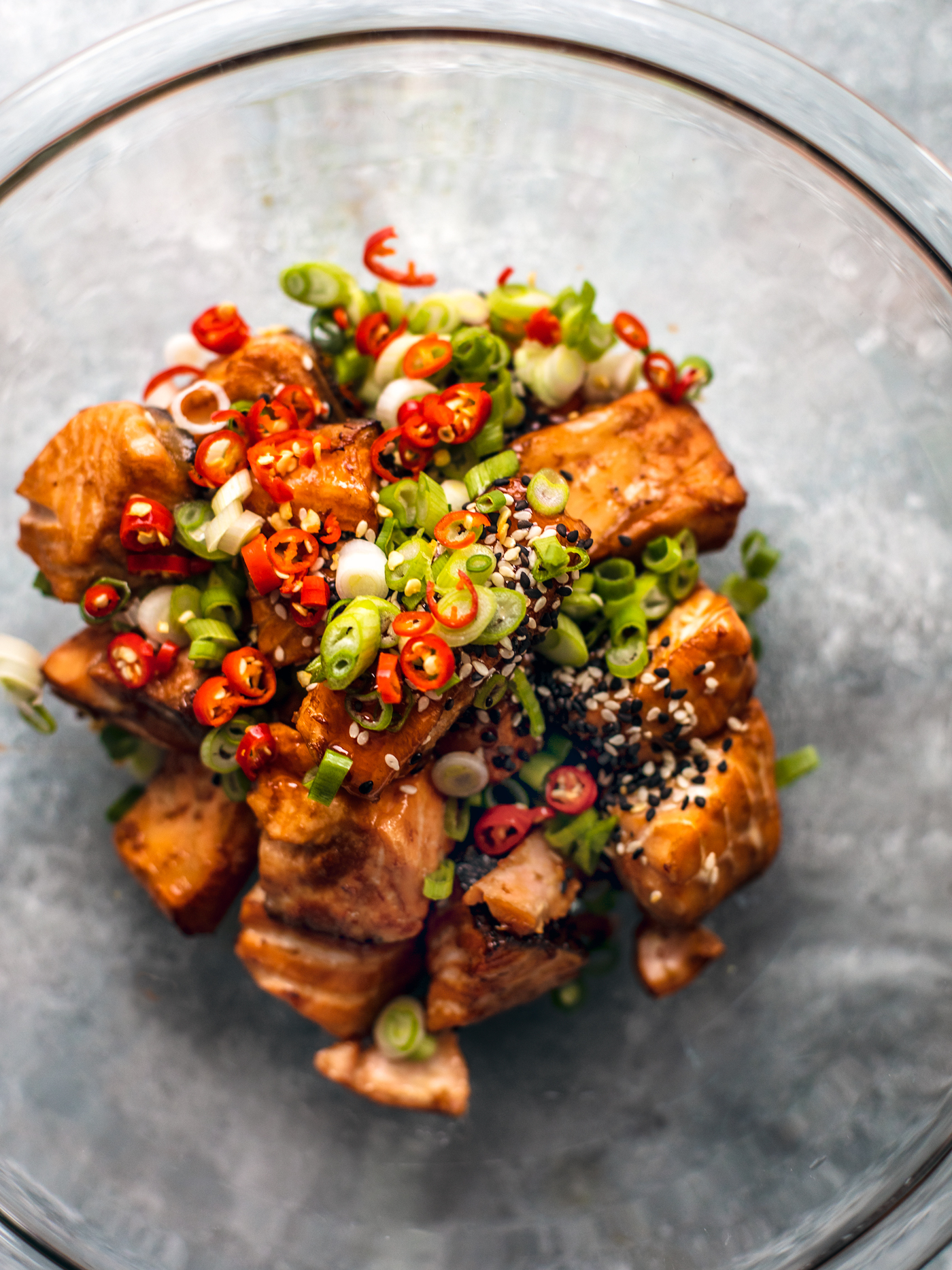 Glass mixing bowl full of salmon bites topped with sweet chili sauce, scallions, sesame seeds, and Thai chilies.