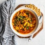 Bowl of vegetable quinoa soup with a spoon, crackers, and striped napkin.