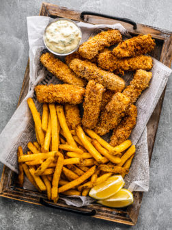 Wooden platter covered in newspaper topped with crispy fish sticks, fries, and tartar sauce.