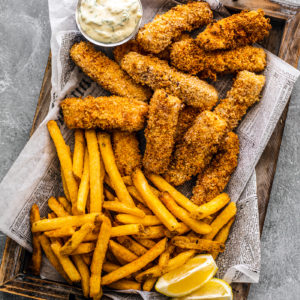 Wooden platter covered in newspaper topped with crispy fish sticks, fries, and tartar sauce.