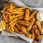 Wooden platter covered in newspaper topped with crispy fish sticks, fries, and tartar sauce.