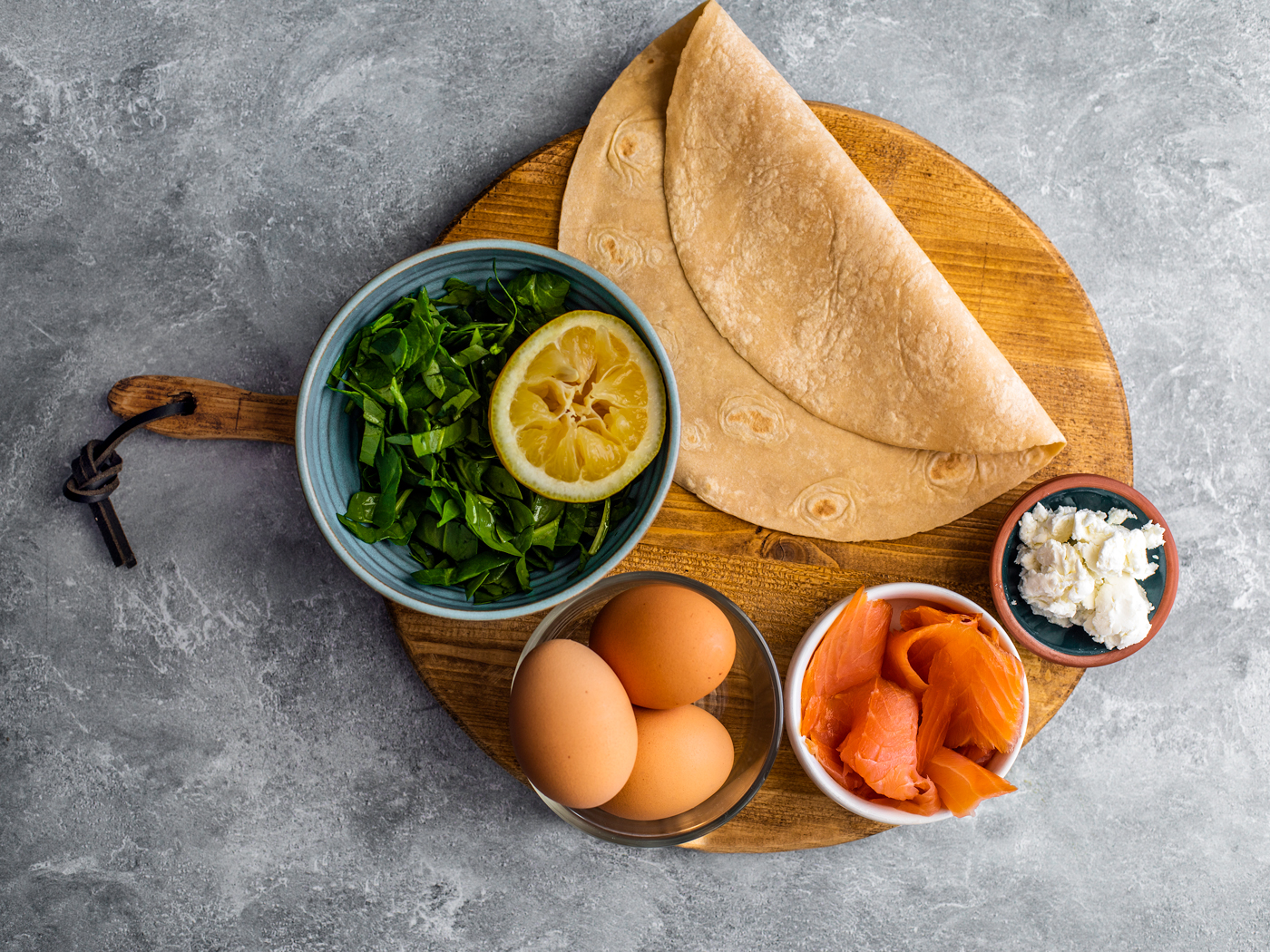 Round serving board with a folded wrap, bowl of baby spinach, eggs, smoked salmon, and goat cheese.