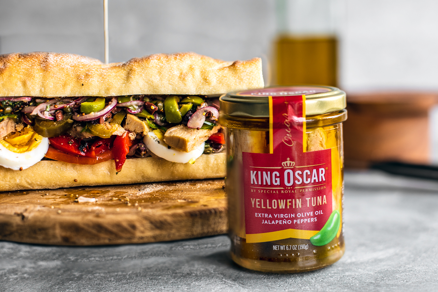 King Oscar jar of tuna standing in front of a big and colorful pan bagnat sandwich.