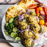 Hands holding out a serving platter of Greek meatballs with fresh veggies, pita, and tzatziki.