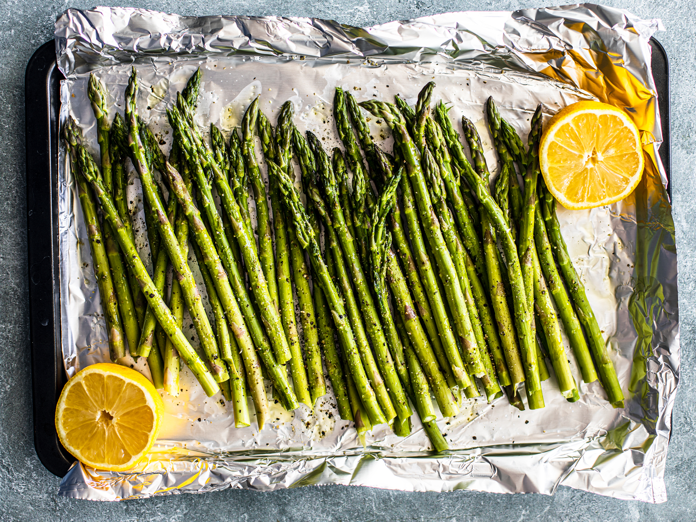 Raw and seasoned asparagus spears on tin foil with lemon halves.