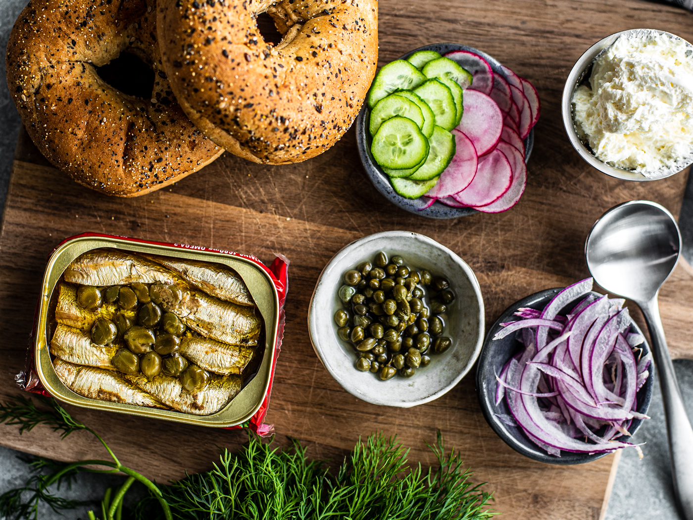 Wooden serving board with bagels, tin of sardines, and bowls of capers, cucumbers, onions, radishes, and cream cheese.