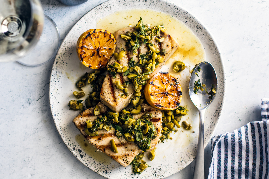 Plate of grilled swordfish with charred lemons and herbs.