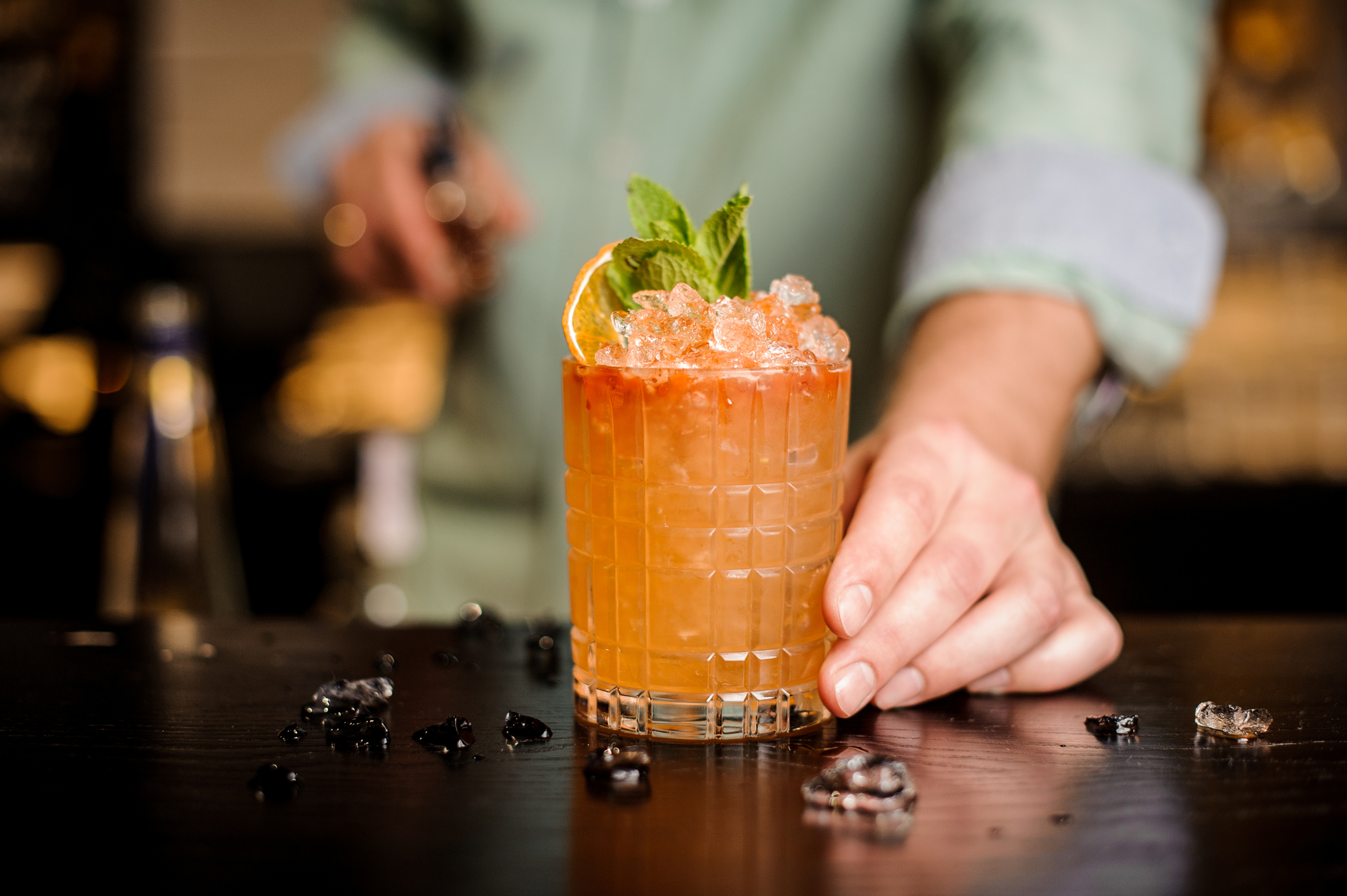 An orange cocktail being presented with an orange slice and mint leaves.