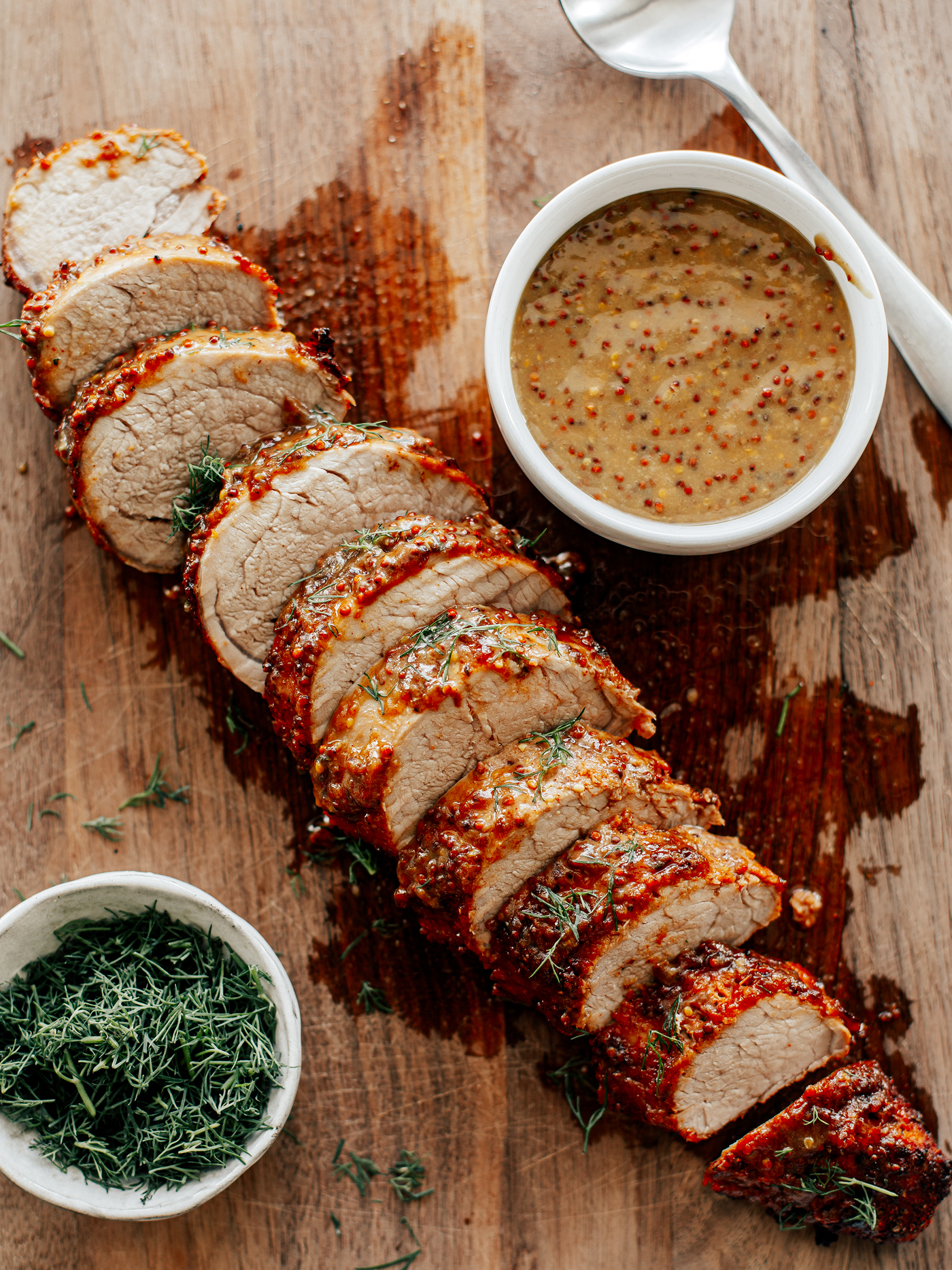 Sliced pork tenderloin on a wooden board with a bowl of mustard sauce and pinch bowl of fresh dill.