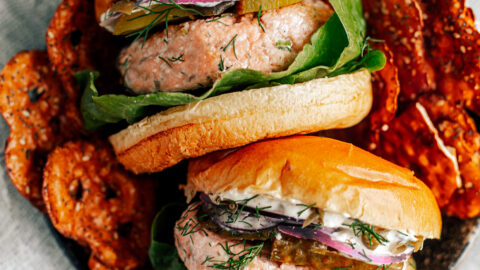 Two salmon burgers in a serving dish stacked with lettuce, pickles, onions, and mayo, with side of pretzels.