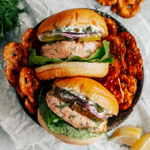 Two salmon burgers in a serving dish stacked with lettuce, pickles, onions, and mayo, with side of pretzels.