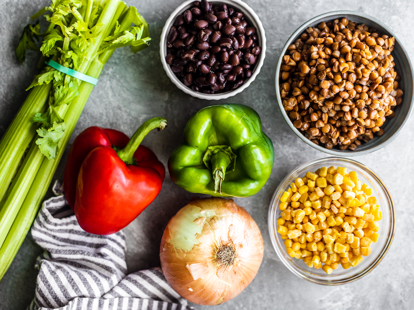 Celery, red bell pepper, green bell pepper, onion, and bowls of black beans, lentils, and corn on a gray background.
