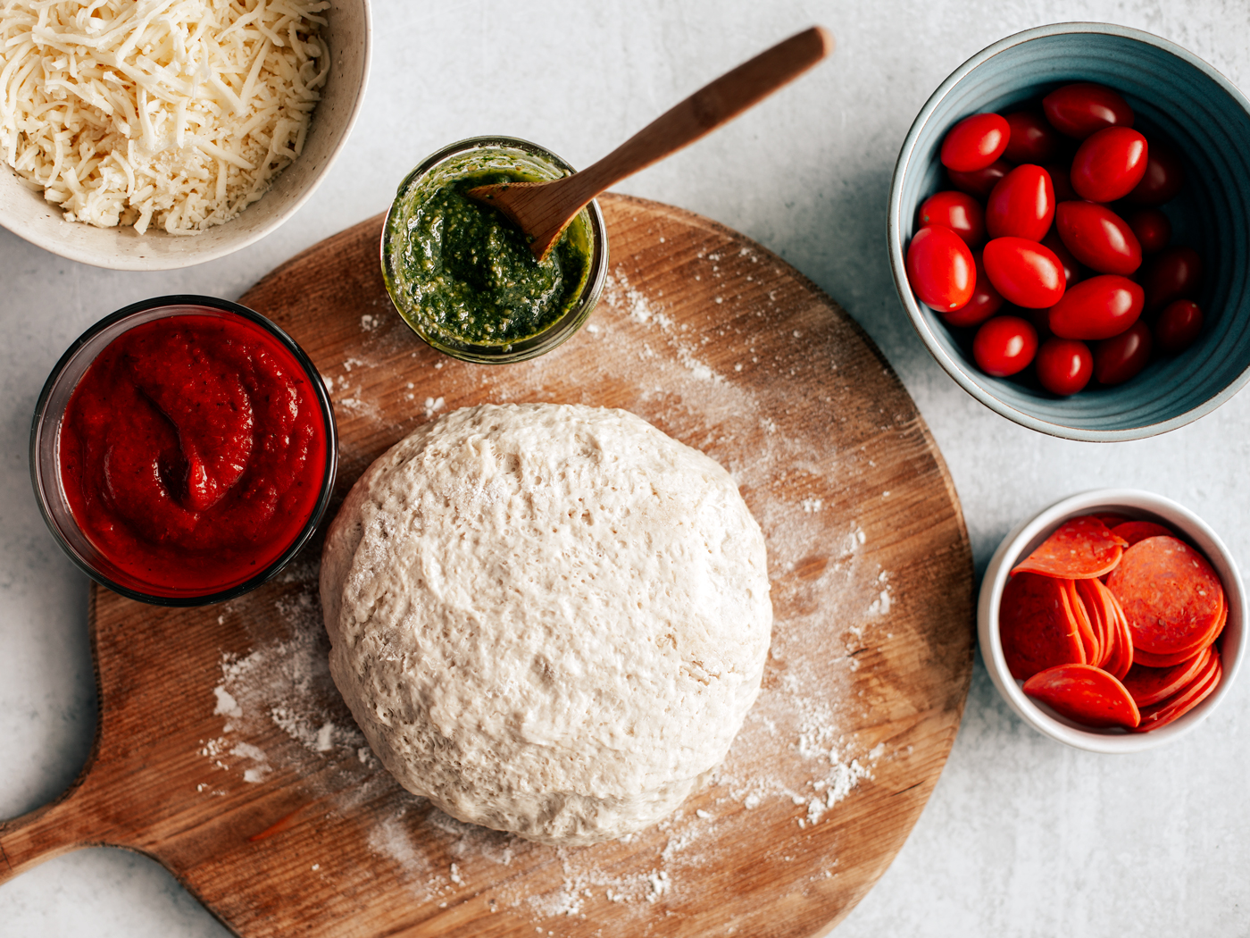 Ball of fresh pizza dough on top of floured wooden cutting board with a bowl of shredded cheese and small jars of sauce, pesto, tomatoes, and pepperoni.