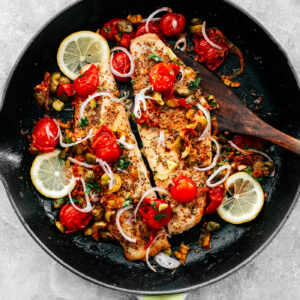 Frying pan with grouper fillets topped with tomatoes, onions, olives, and capers.