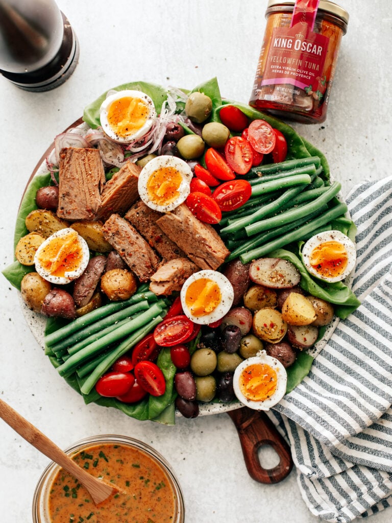 Colorful platter of Ni&ccedil;oise Salad with fresh veggie and soft-boiled eggs. Striped dish towel off to the side with bowl of vinaigrette and jar of tuna.