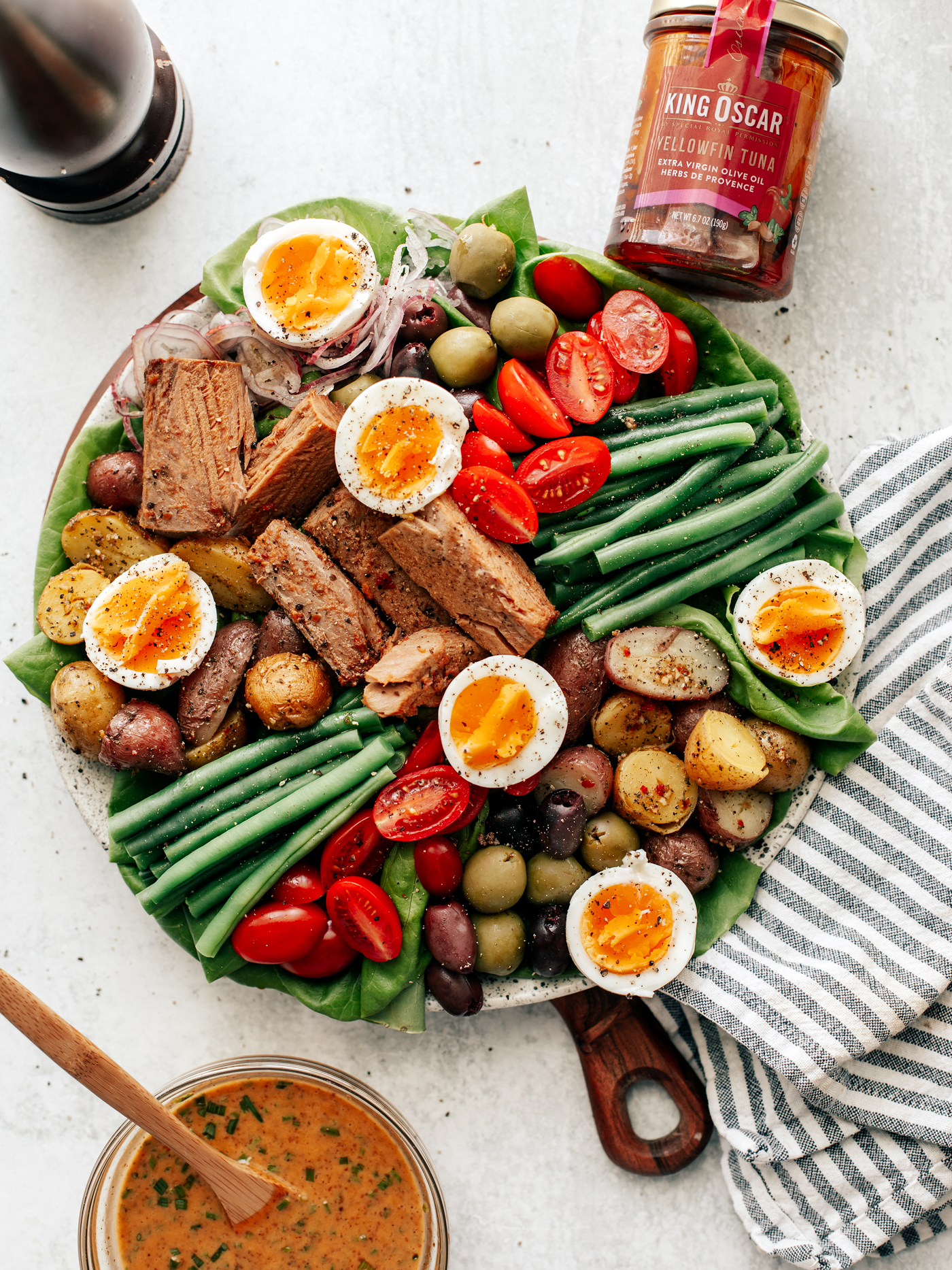Colorful platter of Niçoise Salad with fresh veggie and soft-boiled eggs. Striped dish towel off to the side with bowl of vinaigrette and jar of tuna.