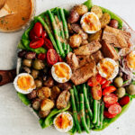 Colorful platter of Niçoise Salad with fresh veggie and soft-boiled egg with bowl of vinaigrette next to it.