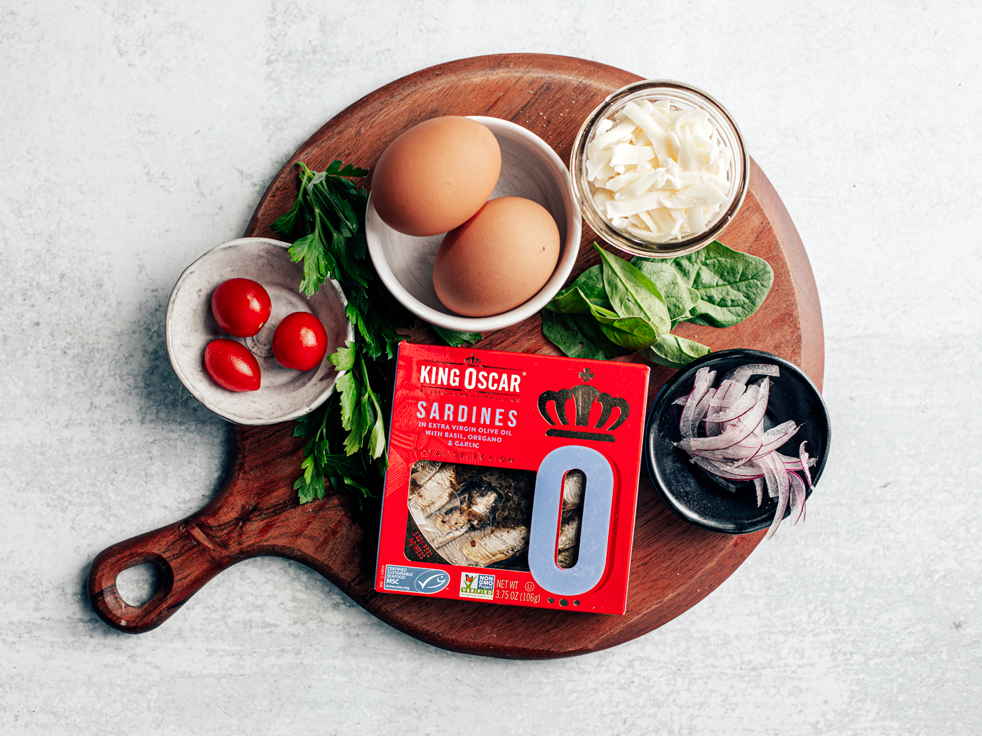 Round wooden serving board with eggs, cherry tomatoes, parsley, spinach, onions, and package of sardines on top.