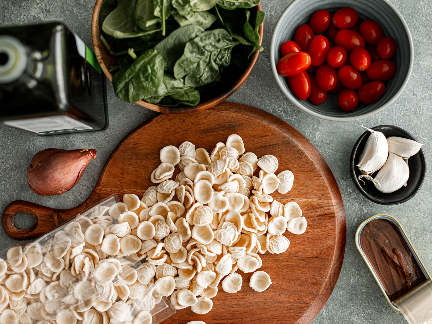 Uncooked orecchiette pasta on cutting board with bowls of spinach, tomatoes, garlic, and a tin of anchovies.