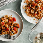 Two white plates of orecchiette pasta with a glass of white wine.