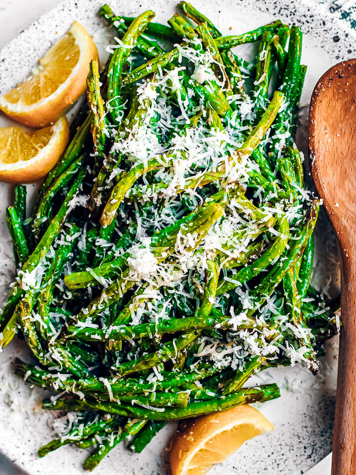 Close up of green beans garnished with parmesan cheese on white serving platter with lemon wedges.