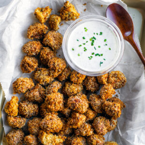 Baking sheet covered with parchment paper and breaded mushrooms with a bowl of ranch.