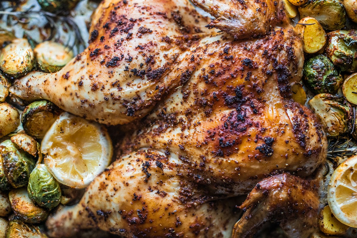 Crispy seasoned spatchcocked chicken on baking sheet with veggies and lemons.