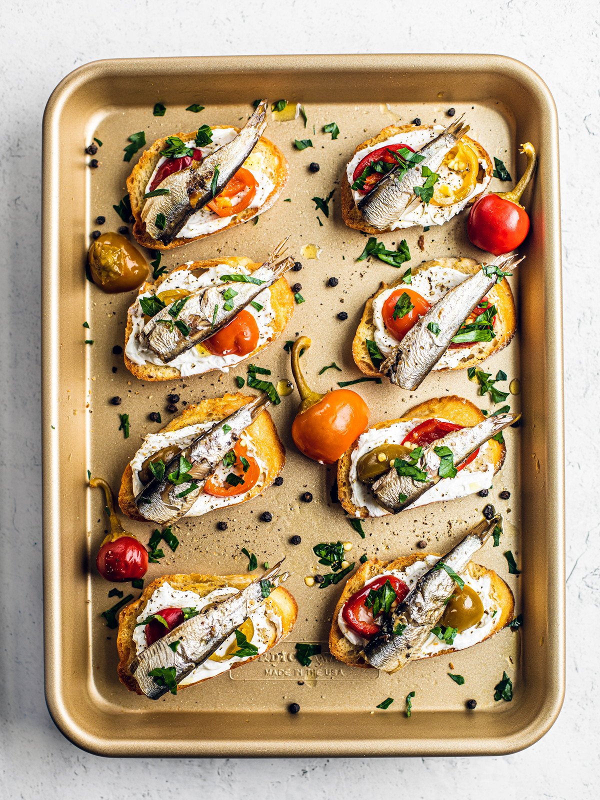 Golden sheet pan with crostini on it, which are topped with goat cheese, cherry peppers, and sardines.