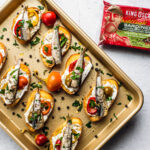Golden sheet pan with crostini on it, which are topped with goat cheese, cherry peppers, and sardines next to a package of sardines.