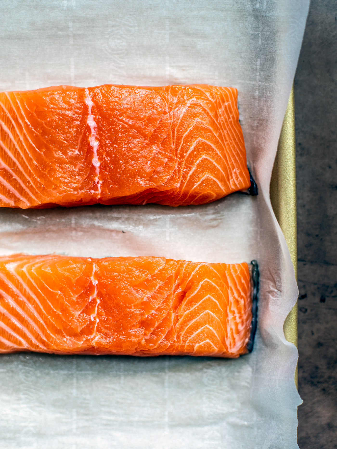 Two bright orange raw fillets of salmon on parchment-covered baking sheet.