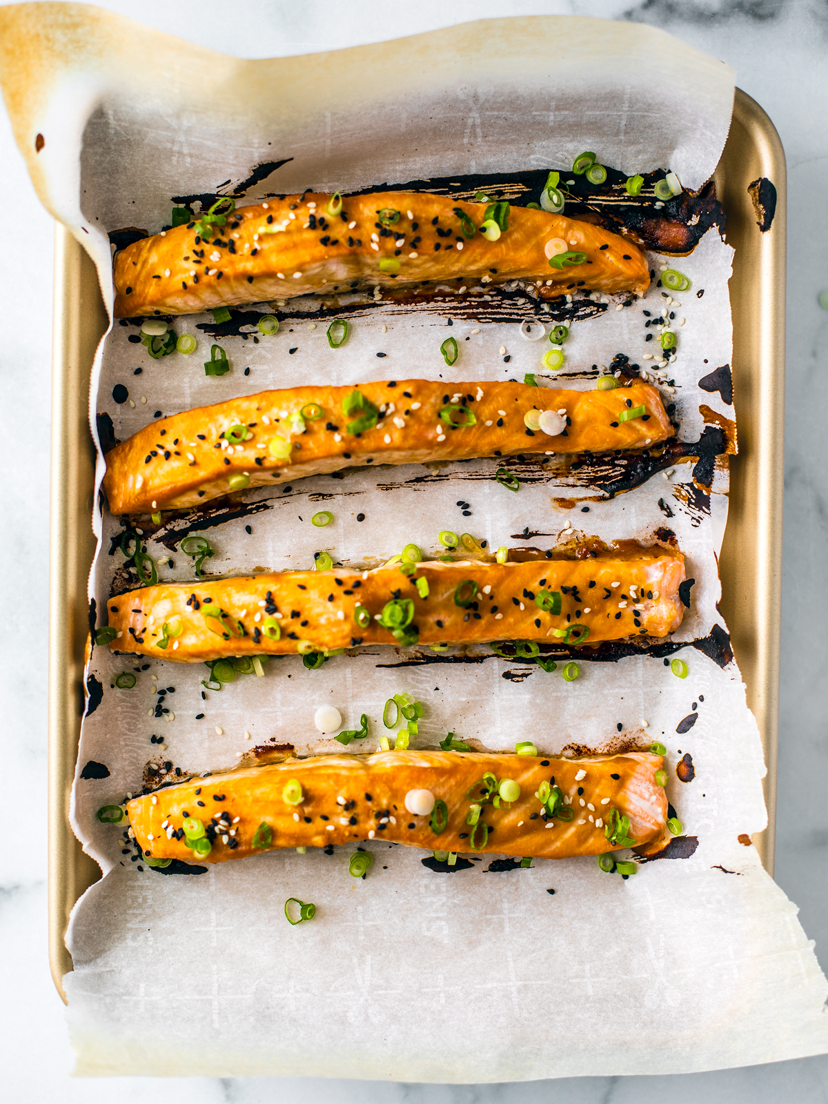 Four salmon filets on a baking sheet with parchment, glazed and garnished with sesame seeds and scallions.