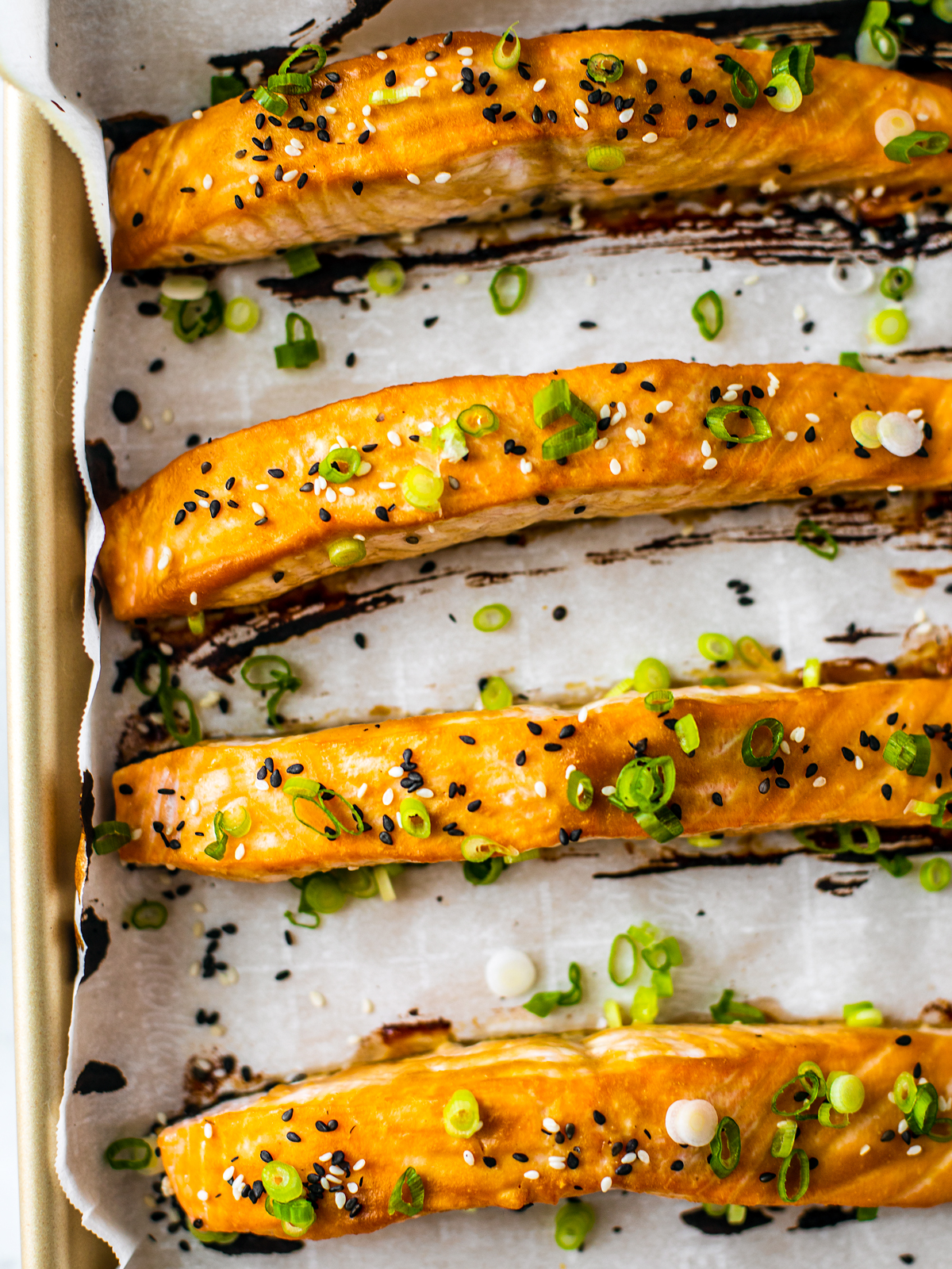Four salmon filets on a baking sheet with parchment, glazed and garnished with sesame seeds and scallions.