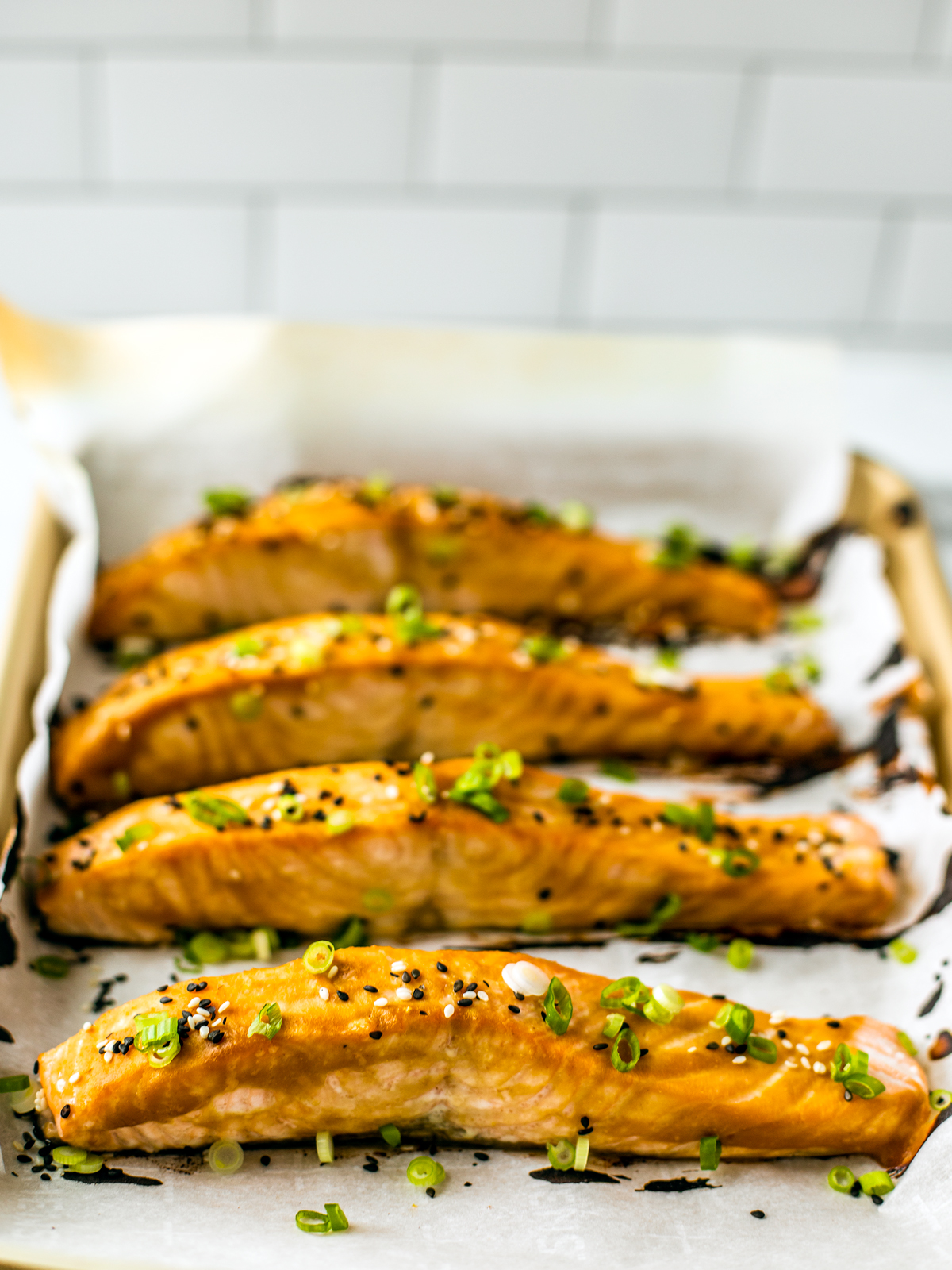 Four salmon filets on a baking sheet with parchment, glazed and garnished with sesame seeds and scallions; a white tile backsplash in the background.
